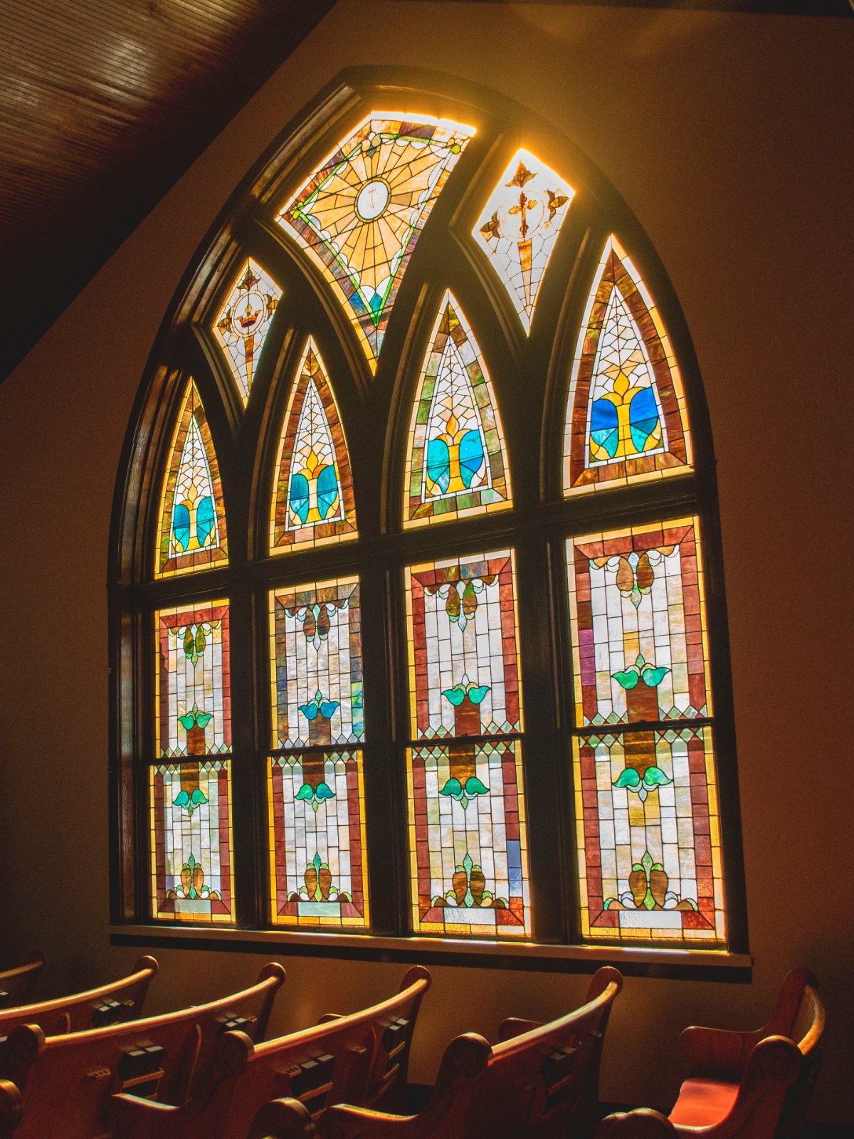 A tall stained glass window in a dimly lit church, wooden pews in the foreground